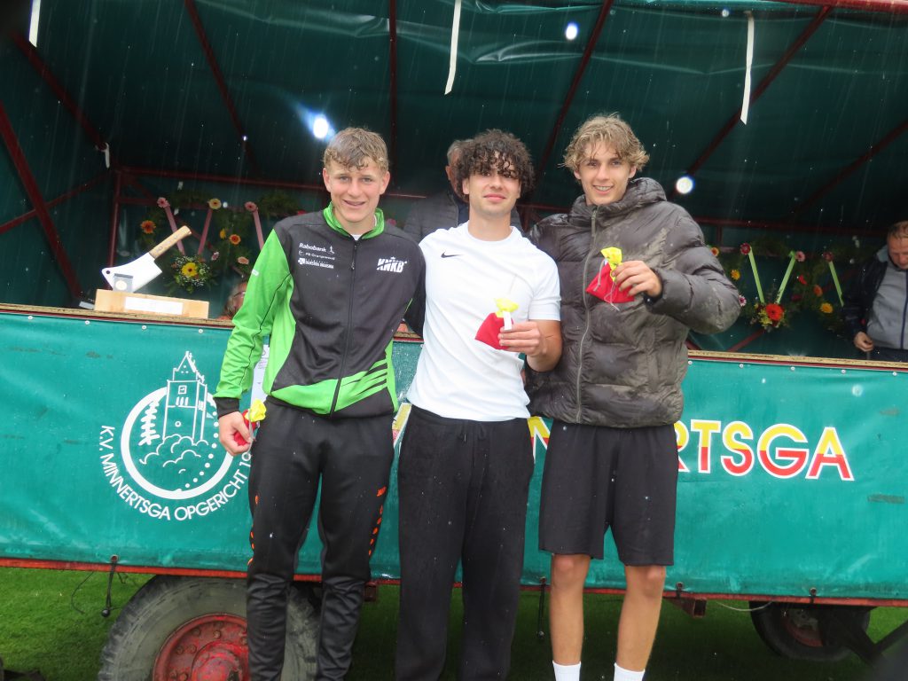 Minnertsga heren 2e klasse vf tweede prijs Karste Hoekstra Tymen Bijlsdma Silvan Elzinga - kaatsnieuws.com Three young men pose on a podium after a race, holding yellow prize bags under a green canopy in light rain.