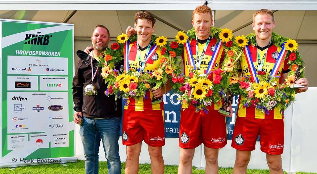 Four cyclists in yellow-and-red kits with flower wreaths and medals pose on a podium with sponsor signage nearby.