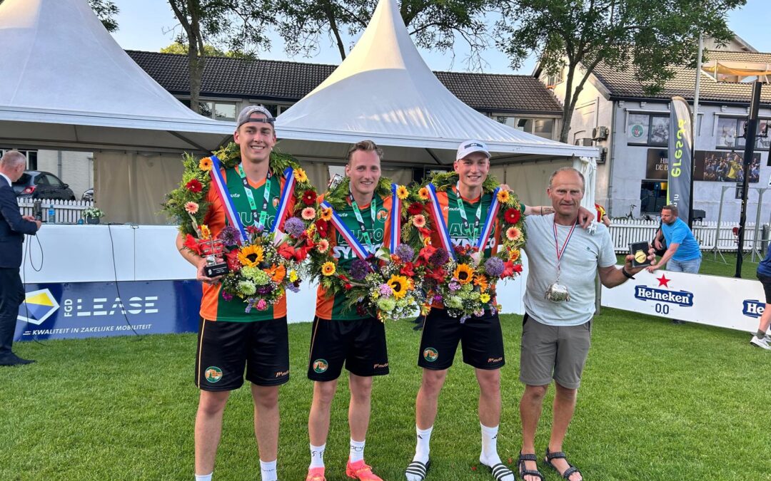 Three young male runners in orange and green uniforms pose with large flower garlands and medals, outdoors at a race event.