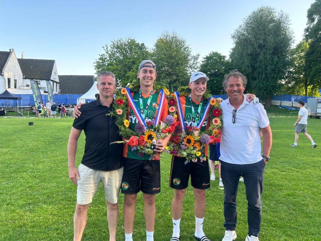 PHOTO-2026-05-25-22-02-41 - kaatsnieuws.com Two athletes in green jerseys with large flower wreaths and medals pose with two men on a sunny grassy field.