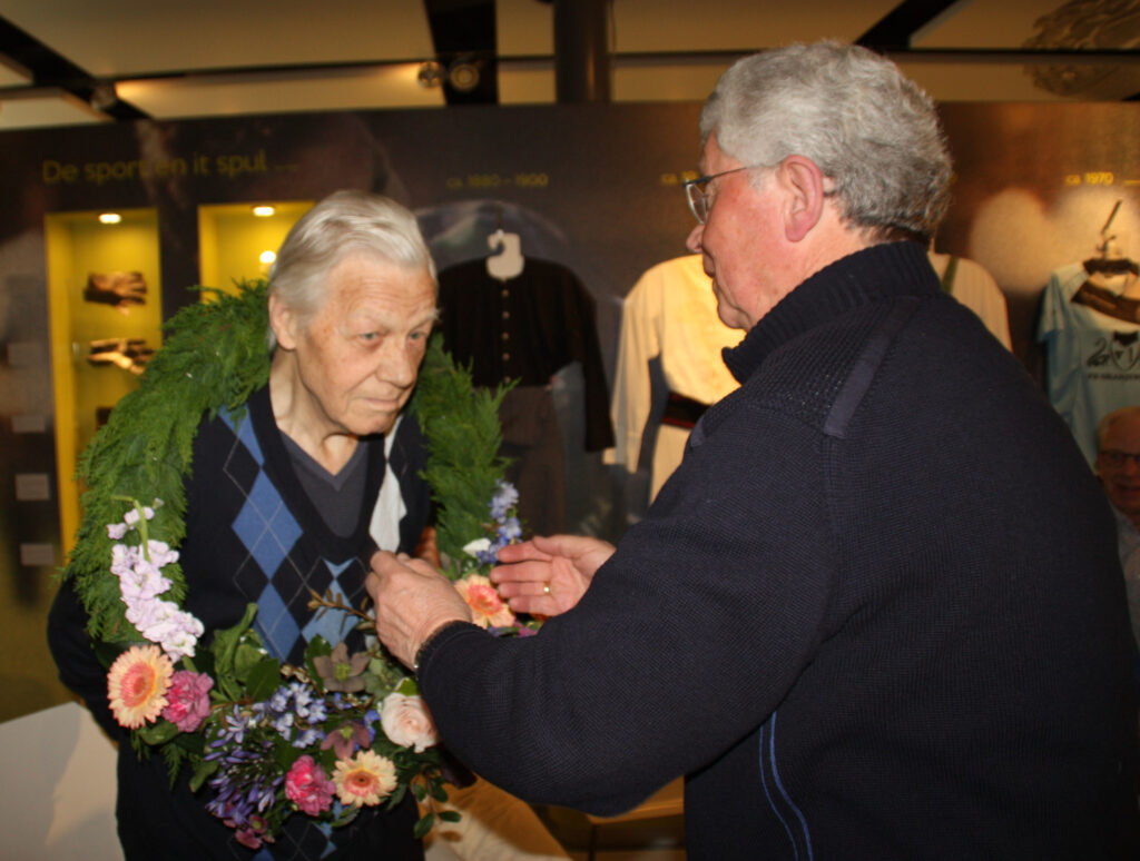 Elderly woman wearing a green flower garland while a man adjusts flowers in an indoor display area.