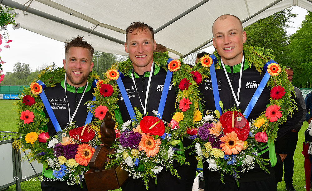 Sint Annaparochie 1 - kaatsnieuws.com Three athletes in black-green uniforms wear medals and blue sashes, posing with large colorful flower wreaths and trophies under a canopy at a sports event.