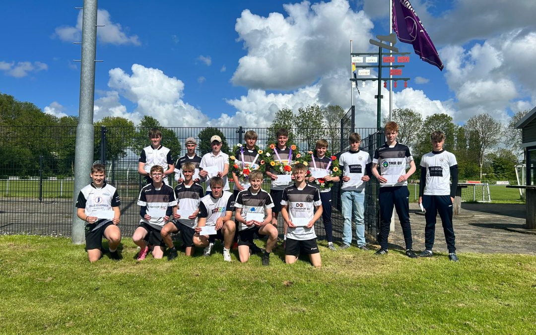 Group of young soccer players posing with flowers and certificates on a grassy field, with a fence and purple flag behind them.