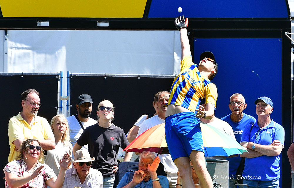 Athlete in yellow-blue uniform leaps to catch a ball in front of an audience at an outdoor event.