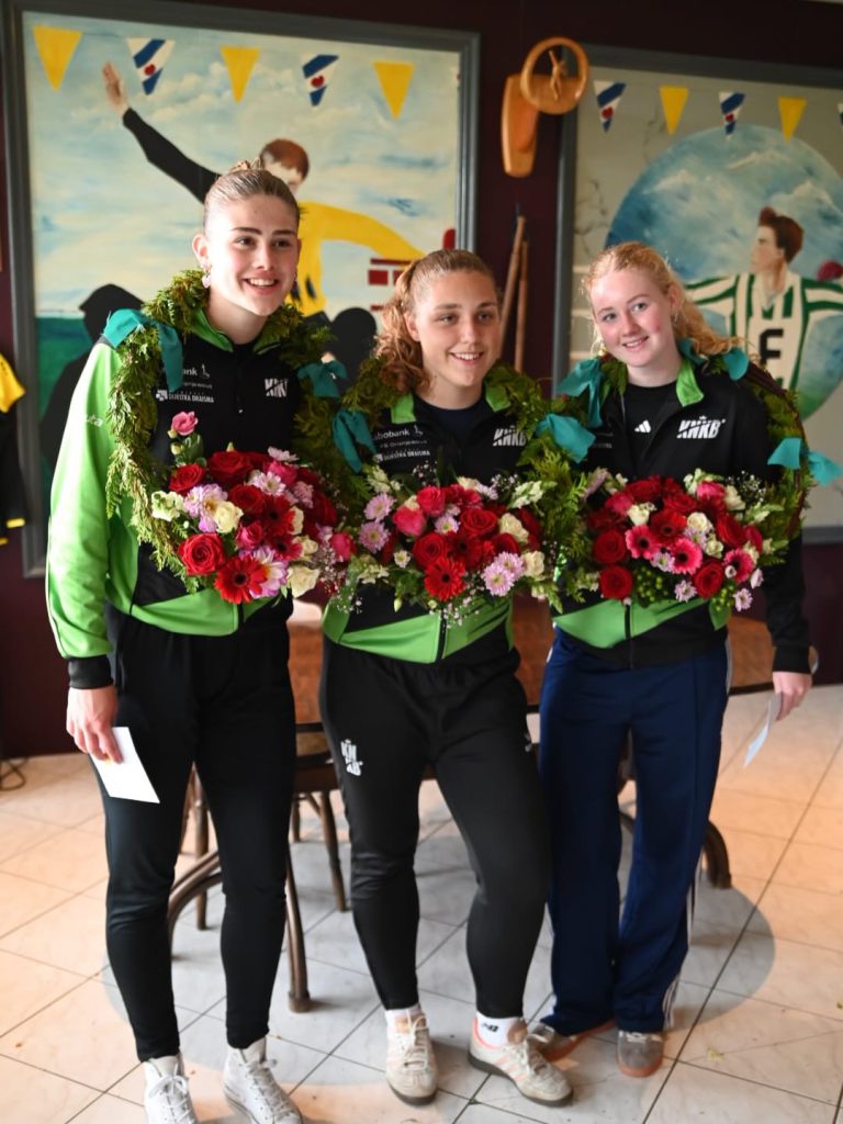 dbroek1 - kaatsnieuws.com Three young female athletes in matching green and black jackets, each with a flower garland around their shoulders, smiling while holding large bouquets of red, pink, and white flowers indoors.