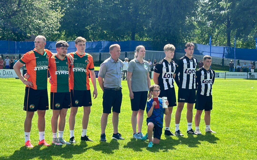 Group of soccer players lined up on a sunny field, wearing orange-green and black-white kits, with spectators in the background.
