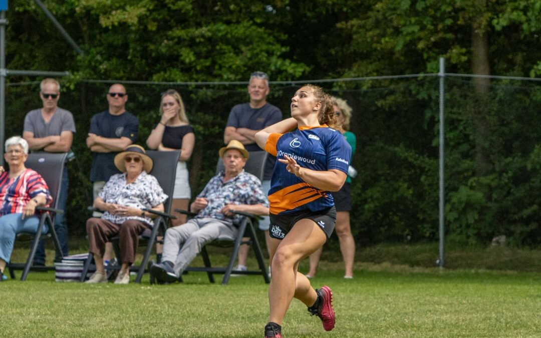 Female runner in a blue-orange jersey sprinting on grass as spectators watch from chairs and standing behind a fence in a park setting.