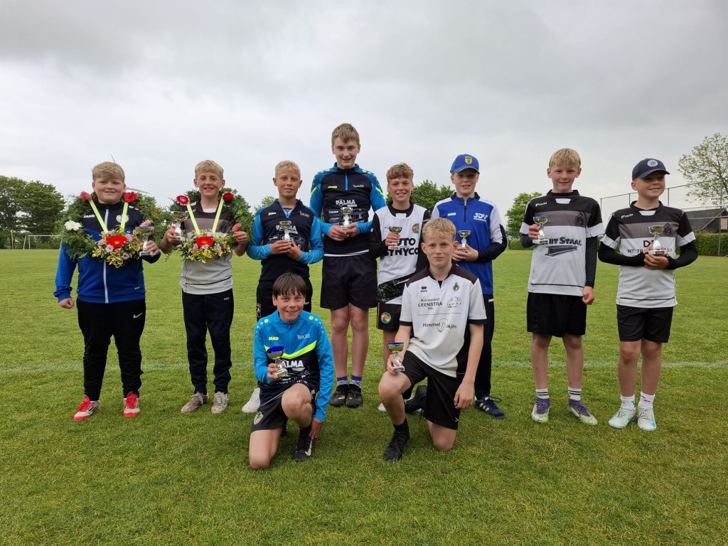 pup b jonges - kaatsnieuws.com Group of young boys posing with trophies on a grass field after a sports event, some holding flowers and medals.
